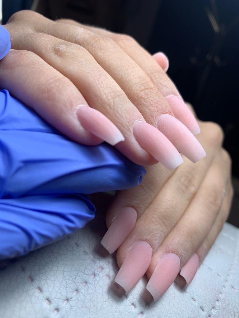 Przybliżenie dłoni z przedłużonymi, kwadratowymi paznokciami w odcieniach jasnego różu i bieli, wykonane przez stylistkę w niebieskich rękawiczkach.