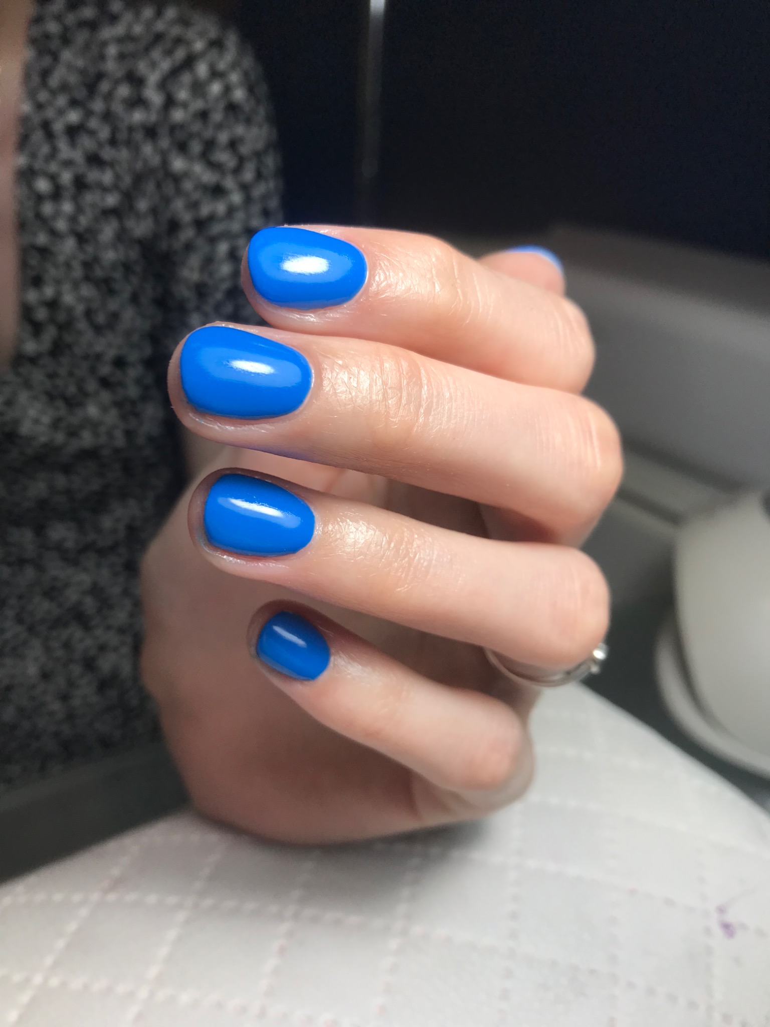 Dłoń z pomalowanymi na jaskrawo niebiesko, krótkimi paznokciami o zaokrąglonym kształcie, na palcu serdecznym srebrny pierścionek z oczkiem, tło rozmyte.