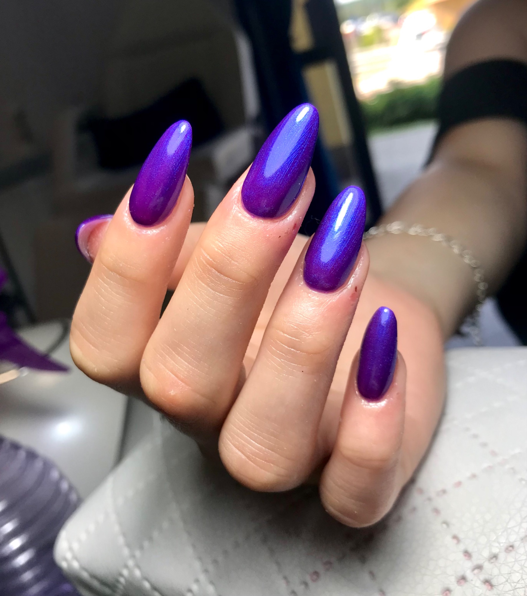 Dłoń z długimi, migdałowymi paznokciami pomalowanymi na fioletowo-niebieski, metaliczny lakier z efektem 'kociego oka', na tle jasnej, pikowanej powierzchni.