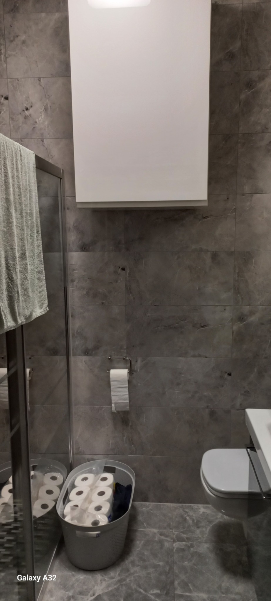 Szara łazienka z marmurowymi płytkami, wisząca szafka nad toaletą, kosz z zapasem papieru toaletowego i fragment kabiny prysznicowej z wiszącym ręcznikiem.