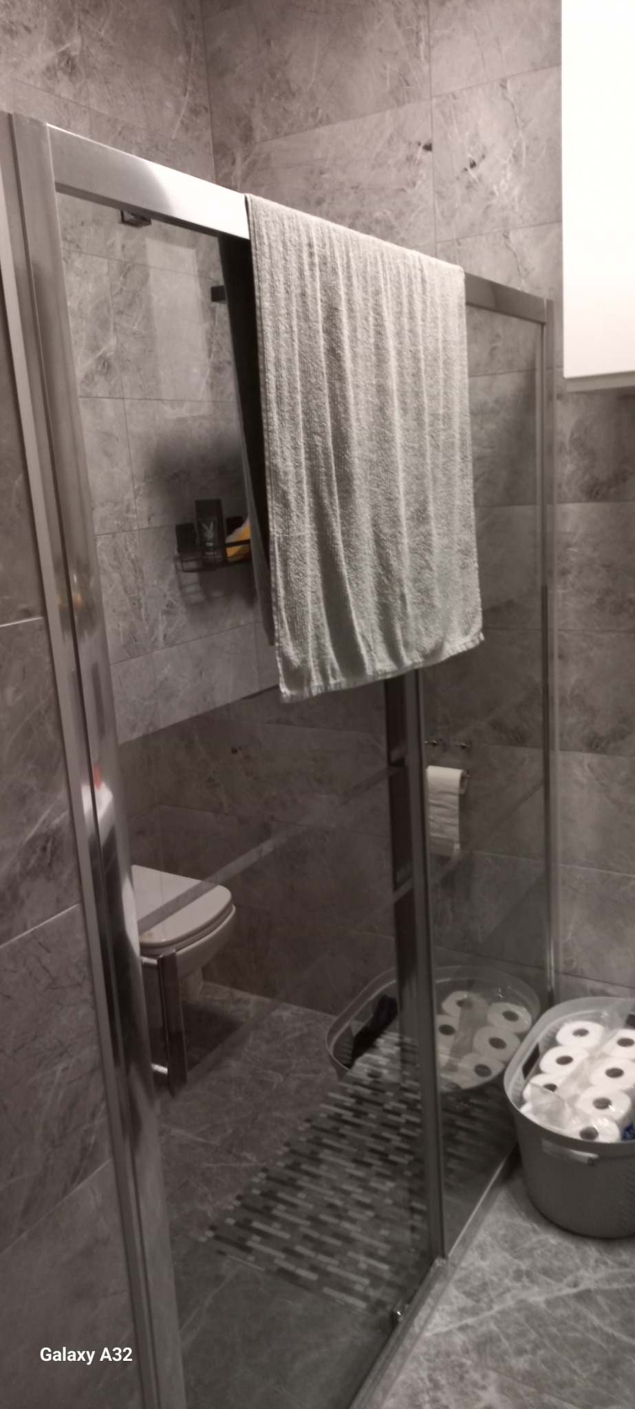 Szklana kabina prysznicowa z szarymi płytkami imitującymi marmur, wiszący ręcznik, toaleta z deską oraz kosz z zapasem papieru toaletowego.