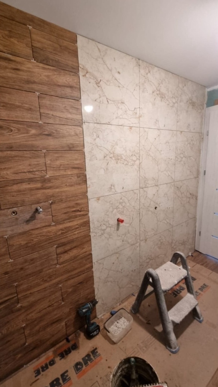 Montaż płytek imitujących drewno i marmur na ścianie łazienki, widoczne narzędzia: wiertarka, drabinka, pojemnik z krzyżykami dystansowymi, na podłodze kartony zabezpieczające.