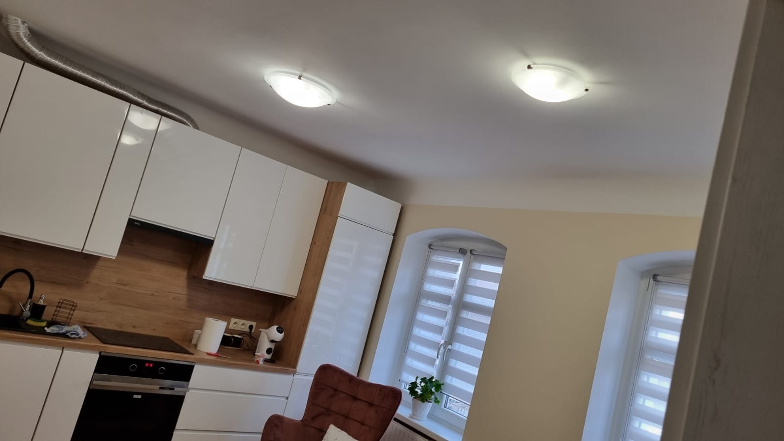 Nowoczesna kuchnia z białymi szafkami, drewnianym panelem ściennym, oknami z roletami wnękowymi i wentylacją ze stali nierdzewnej, oświetlona dwoma okrągłymi lampami sufitowymi.