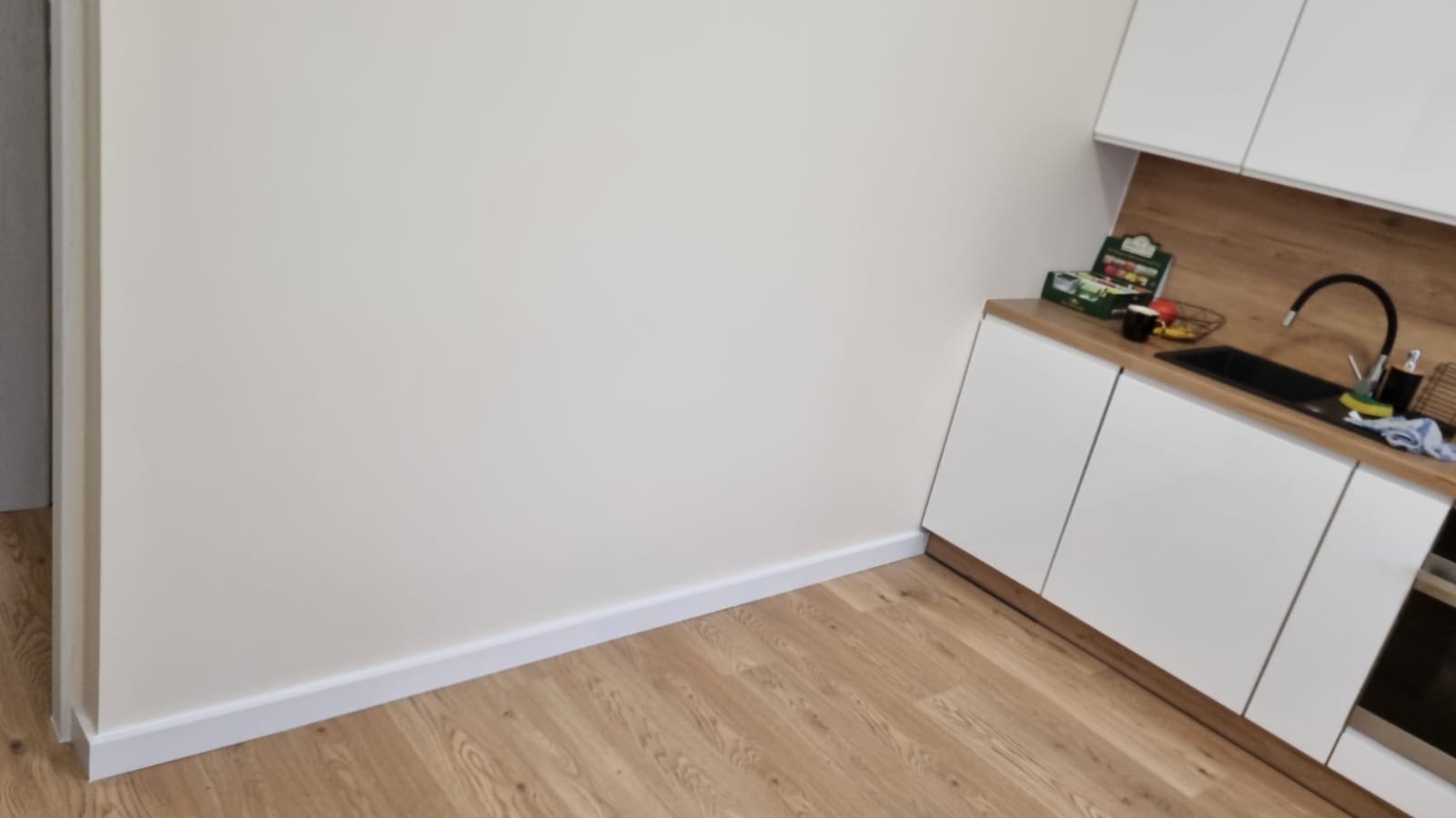 Jasna kuchnia po remoncie: białe szafki, drewniany blat, ciemna armatura, panele podłogowe, świeżo pomalowane ściany z białą listwą przypodłogową.