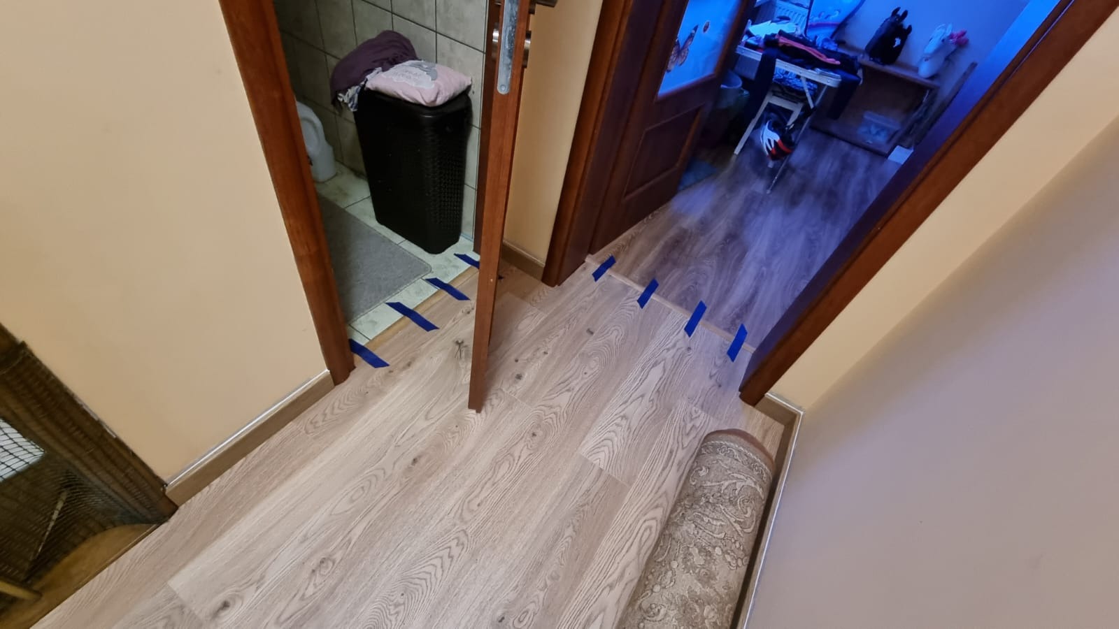 Fragment podłogi z jasnych paneli, zabezpieczonych niebieską taśmą malarską wzdłuż framug drzwi, częściowo zwinięty dywan leżący na podłodze.
