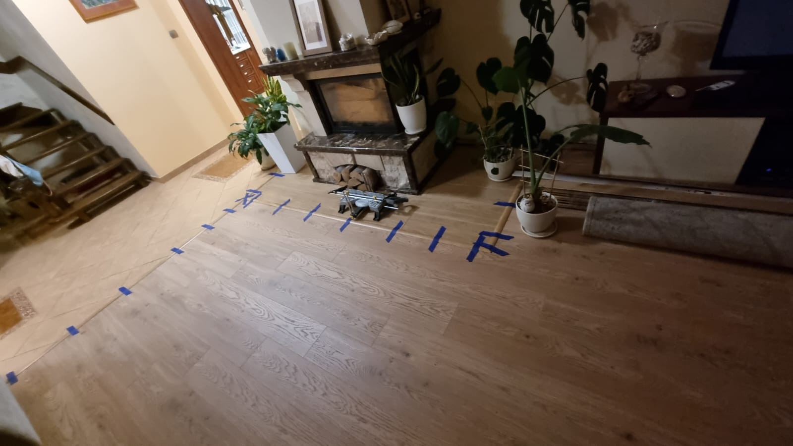 Układanie parkietu w salonie z kominkiem, widoczne linie wyznaczone taśmą malarską, piła do cięcia paneli, rośliny doniczkowe i schody w tle.