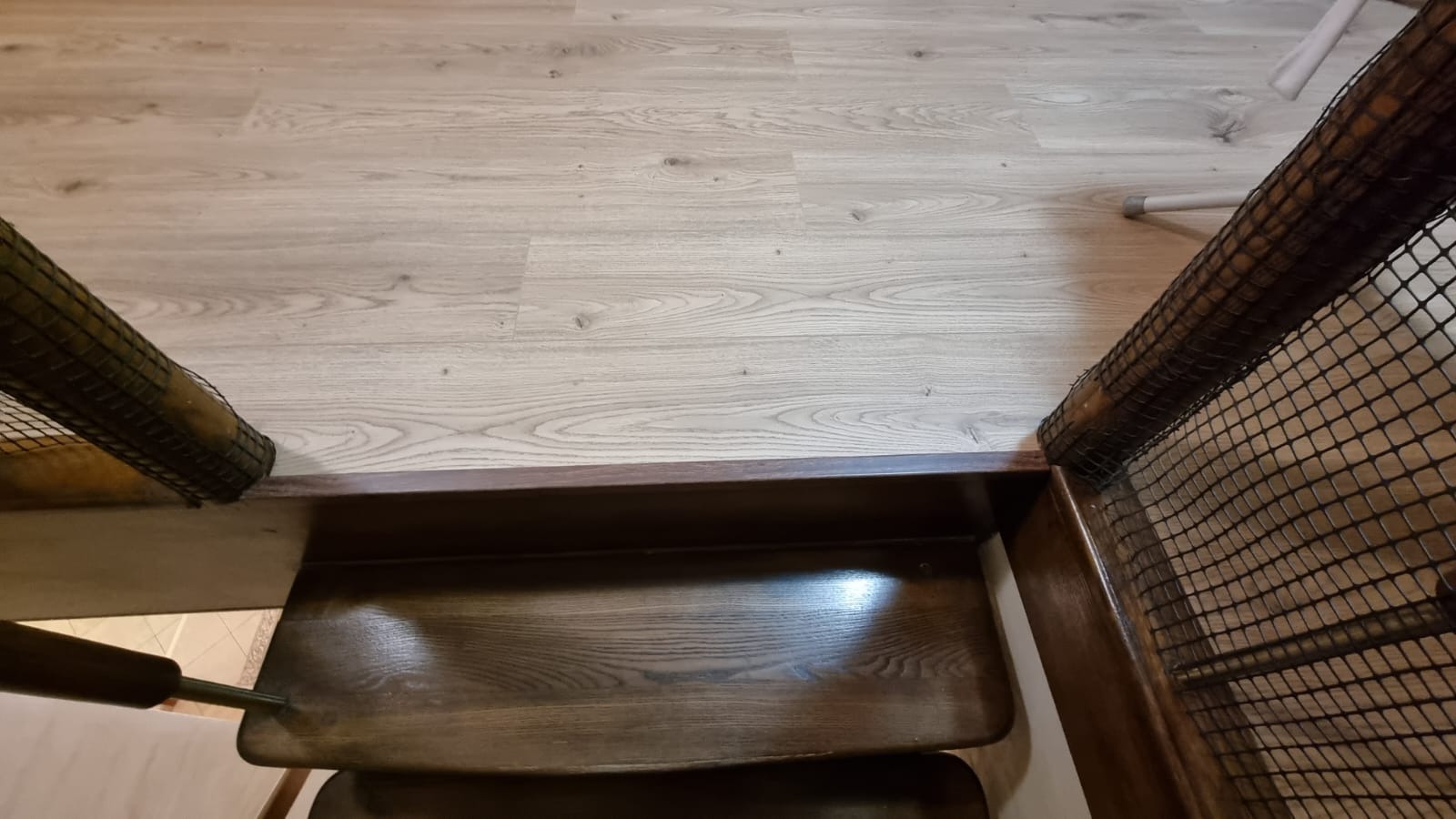 Jasne panele podłogowe sąsiadują z ciemnymi, drewnianymi schodami zabezpieczonymi siatką ochronną.