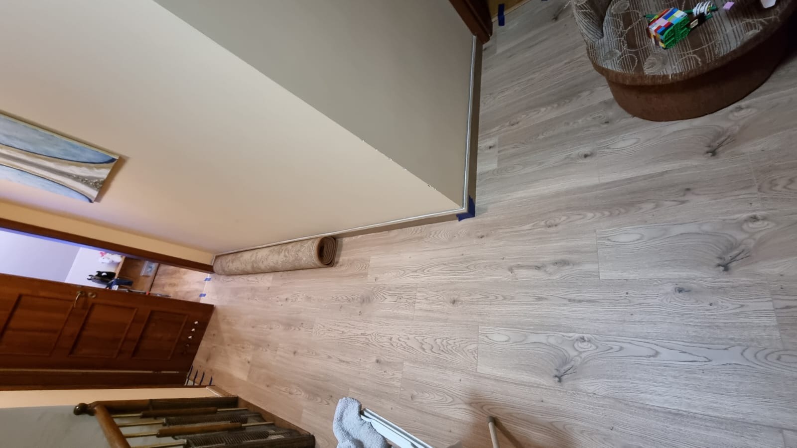 Jasne panele podłogowe z widocznym wzorem drewna, zabezpieczone niebieską taśmą malarską przy ścianie, z roletą zwiniętą w rogu i fragmentem schodów w dolnej części kadru.