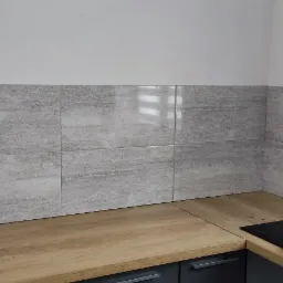 Wykończona ściana nad blatem kuchennym z szarymi, matowymi płytkami imitującymi kamień, drewniany blat, czarna płyta indukcyjna z czajnikiem ze stali nierdzewnej i mikrofalówka w tle.