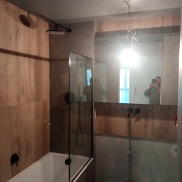 Serhii Pobokov - Nowoczesna łazienka z wanną i szklanym parawanem, drewnopodobne płytki na ścianach, czarna armatura, widoczne odbicie osoby robiącej zdjęcie w lustrze, surowe wykończenie pod umywalką oraz wiadro...