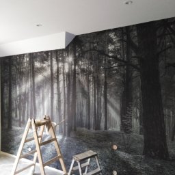 Serhii Pobokov - Fototapeta z motywem leśnym na ścianie w trakcie montażu, widoczne drabiny malarskie i fragment sufitu z oświetleniem punktowym.