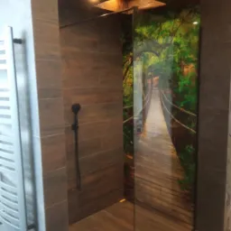 Nowoczesna kabina prysznicowa z panelem szklanym z nadrukiem mostu linowego w dżungli, brązowe płytki imitujące drewno na ścianach, czarna deszczownica, grzejnik drabinkowy.