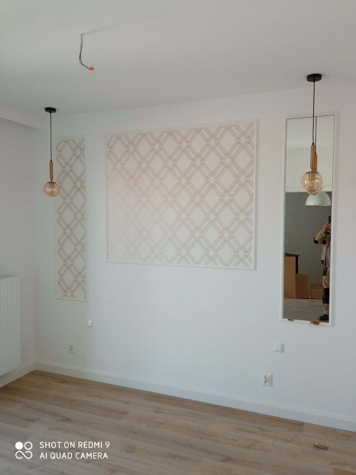 Wykończone wnętrze pokoju z panelami dekoracyjnymi w geometryczny wzór, wiszącymi lampami z okrągłymi kloszami i lustrem w białej ramie, ukazującym odbicie osoby robiącej zdjęcie.
