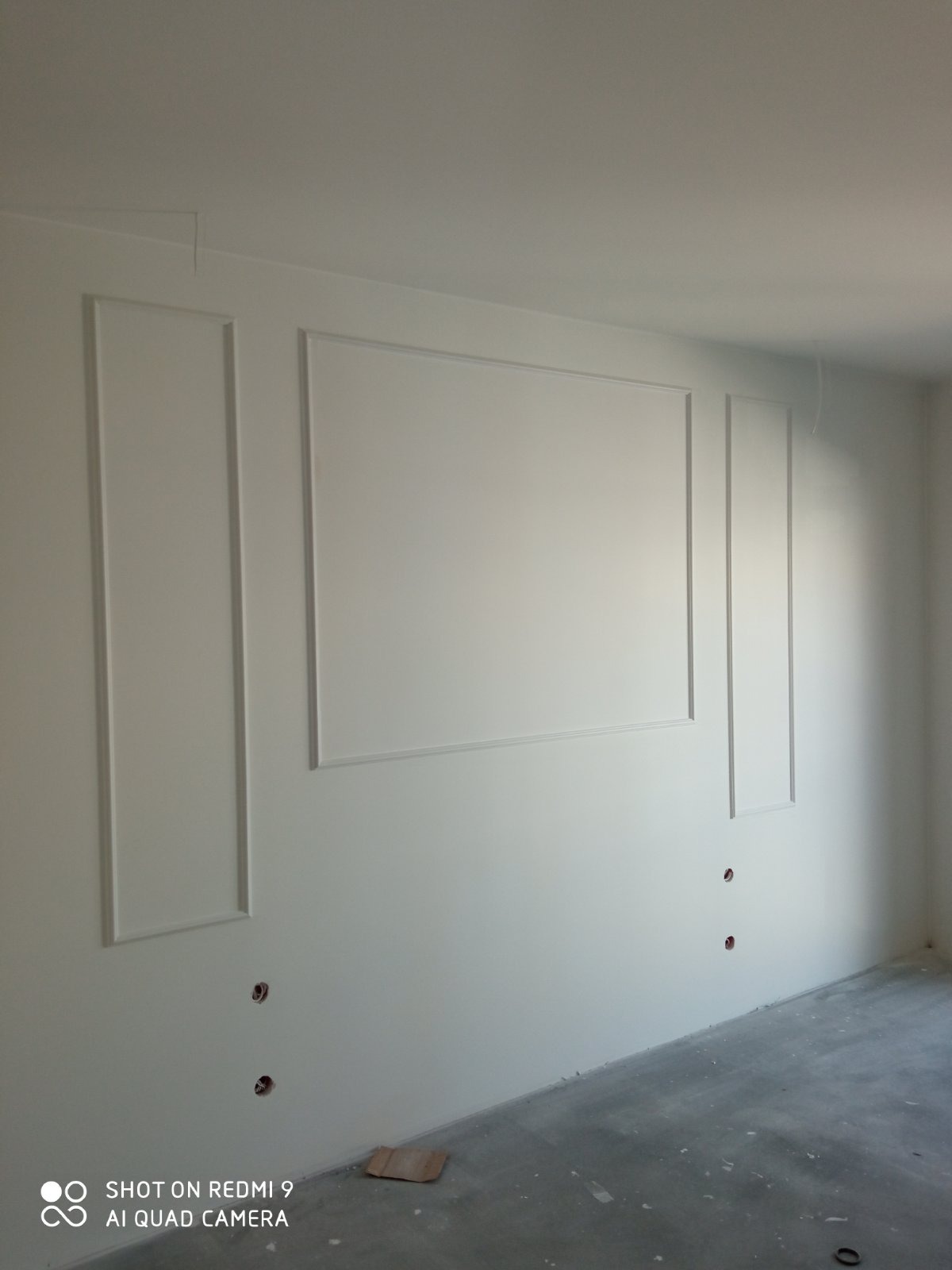 Ściana w pokoju z nowymi, białymi sztukateriami w formie prostokątnych ramek, widoczne puszki elektryczne w ścianie i surowa betonowa podłoga.