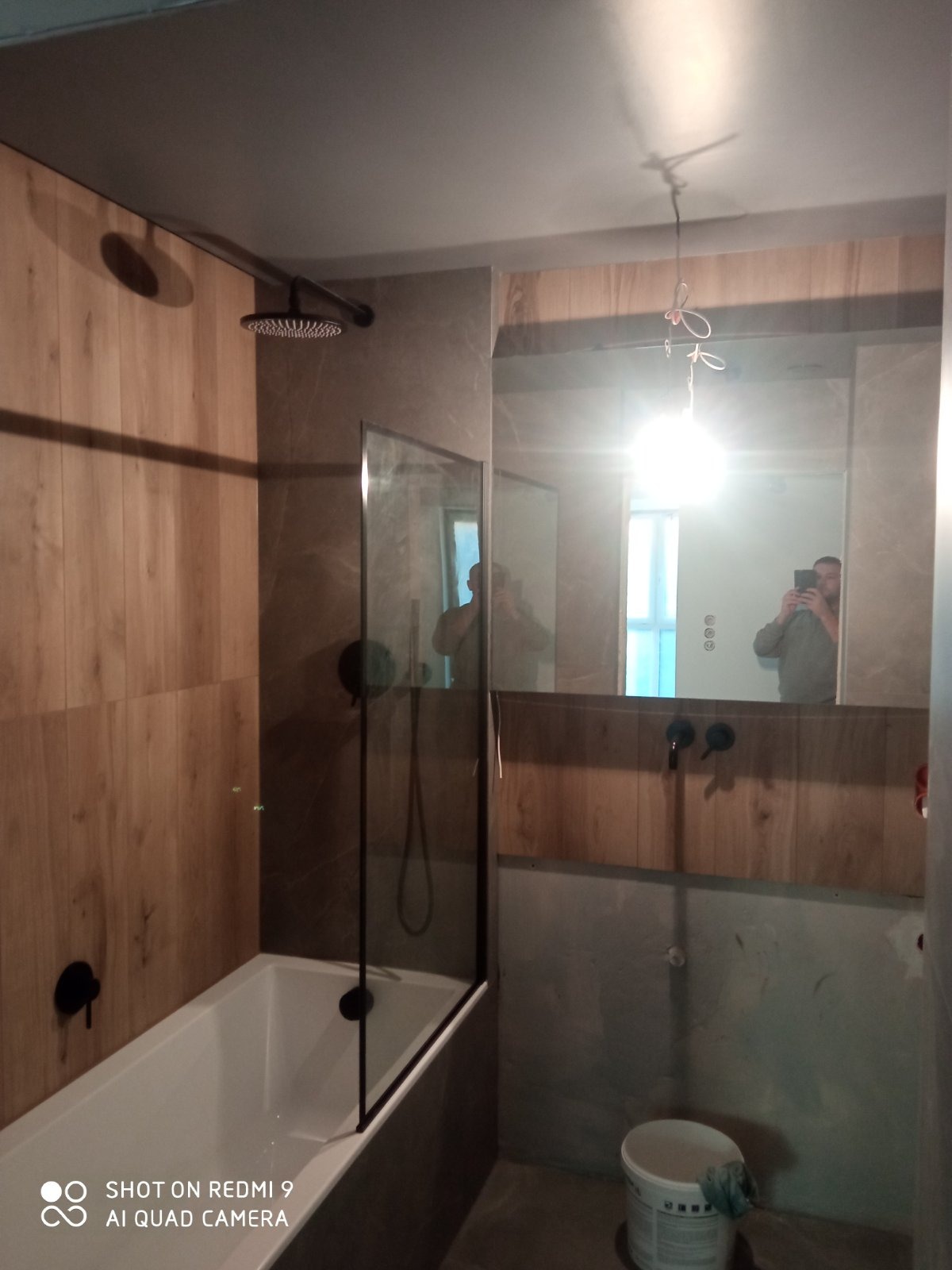 Nowoczesna łazienka z wanną i szklanym parawanem, drewnopodobne płytki na ścianach, czarna armatura, widoczne odbicie osoby robiącej zdjęcie w lustrze, surowe wykończenie pod umywalką oraz wiadro...