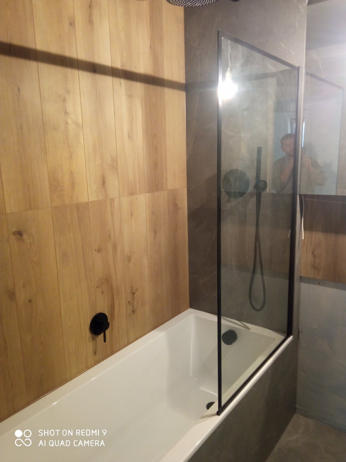 Nowoczesna łazienka z wanną i szklanym parawanem, drewnopodobne płytki na ścianie, czarna armatura, widoczne odbicie osoby robiącej zdjęcie.