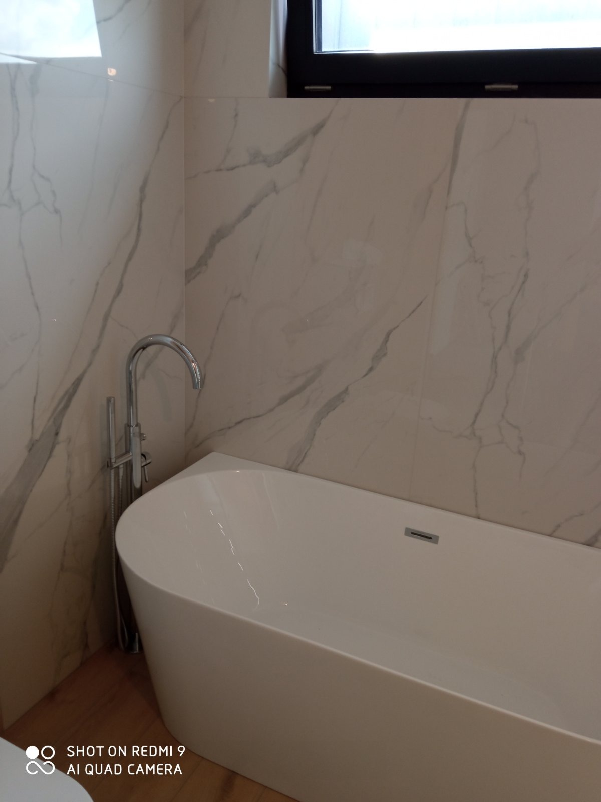 Nowoczesna, wolnostojąca wanna z chromowaną baterią podłogową na tle ściany wyłożonej płytkami imitującymi marmur z czarnym oknem w górnym rogu.