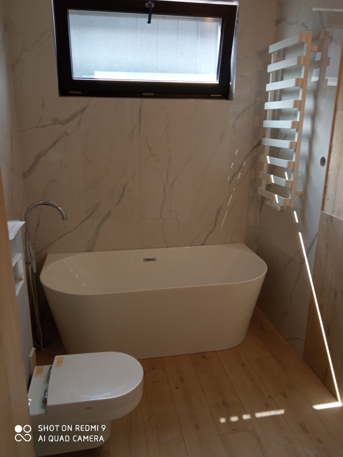 Nowoczesna, jasna łazienka z wolnostojącą wanną, podwieszaną toaletą i grzejnikiem drabinkowym na ścianie z marmurowym wzorem, oświetlona naturalnym światłem z okna.