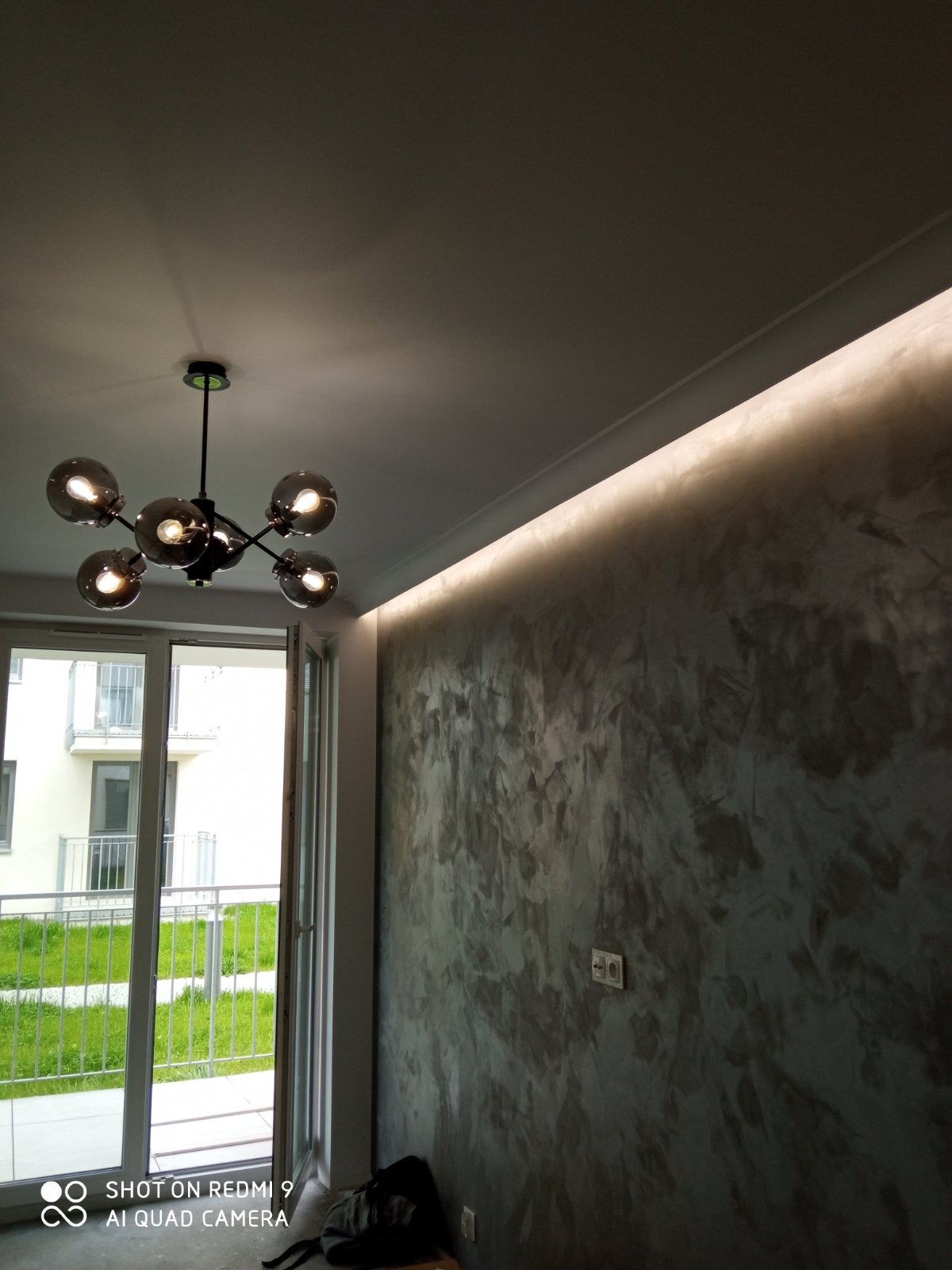 Szara ściana z dekoracyjnym tynkiem, oświetlona listwą LED pod sufitem, nowoczesna lampa sufitowa z kulistymi kloszami, okno balkonowe z widokiem na zieleń.