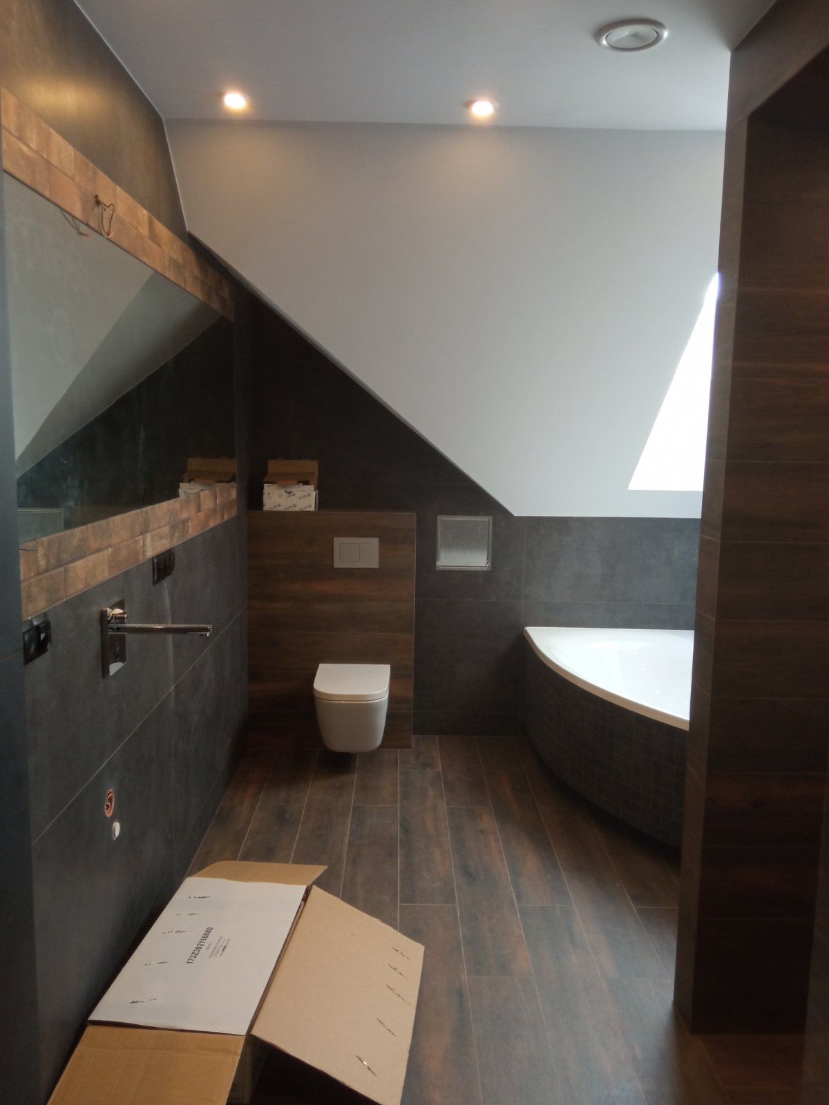 Nowoczesna łazienka na poddaszu z drewnopodobnymi płytkami, wiszącą toaletą i asymetryczną wanną, oświetlona punktowymi lampami, z rozłożonym kartonem na podłodze.