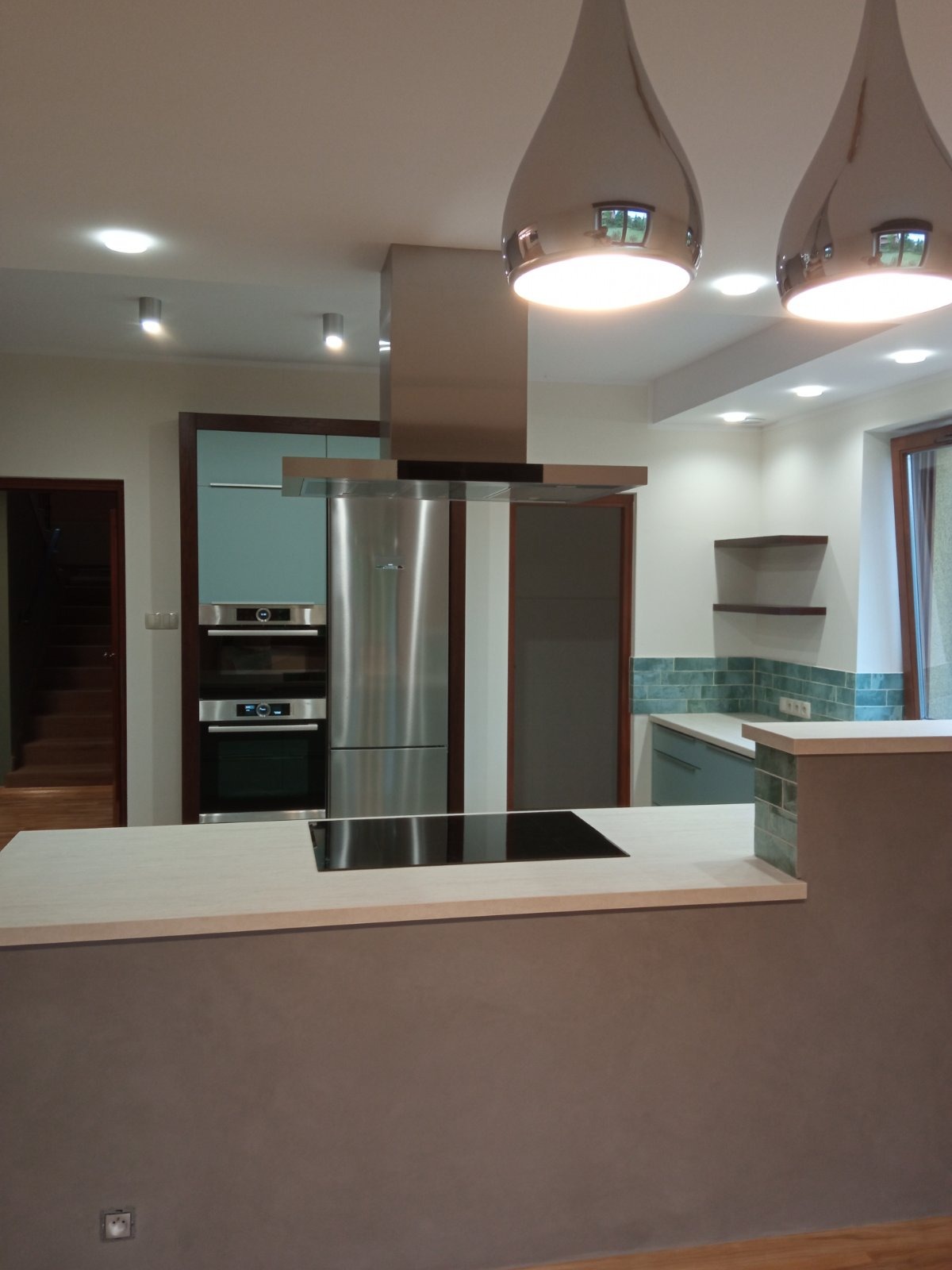 Nowoczesna kuchnia z wyspą, płyta indukcyjna w blacie, zabudowa kuchenna z piekarnikiem i lodówką w stali nierdzewnej, oświetlenie punktowe i wiszące lampy.