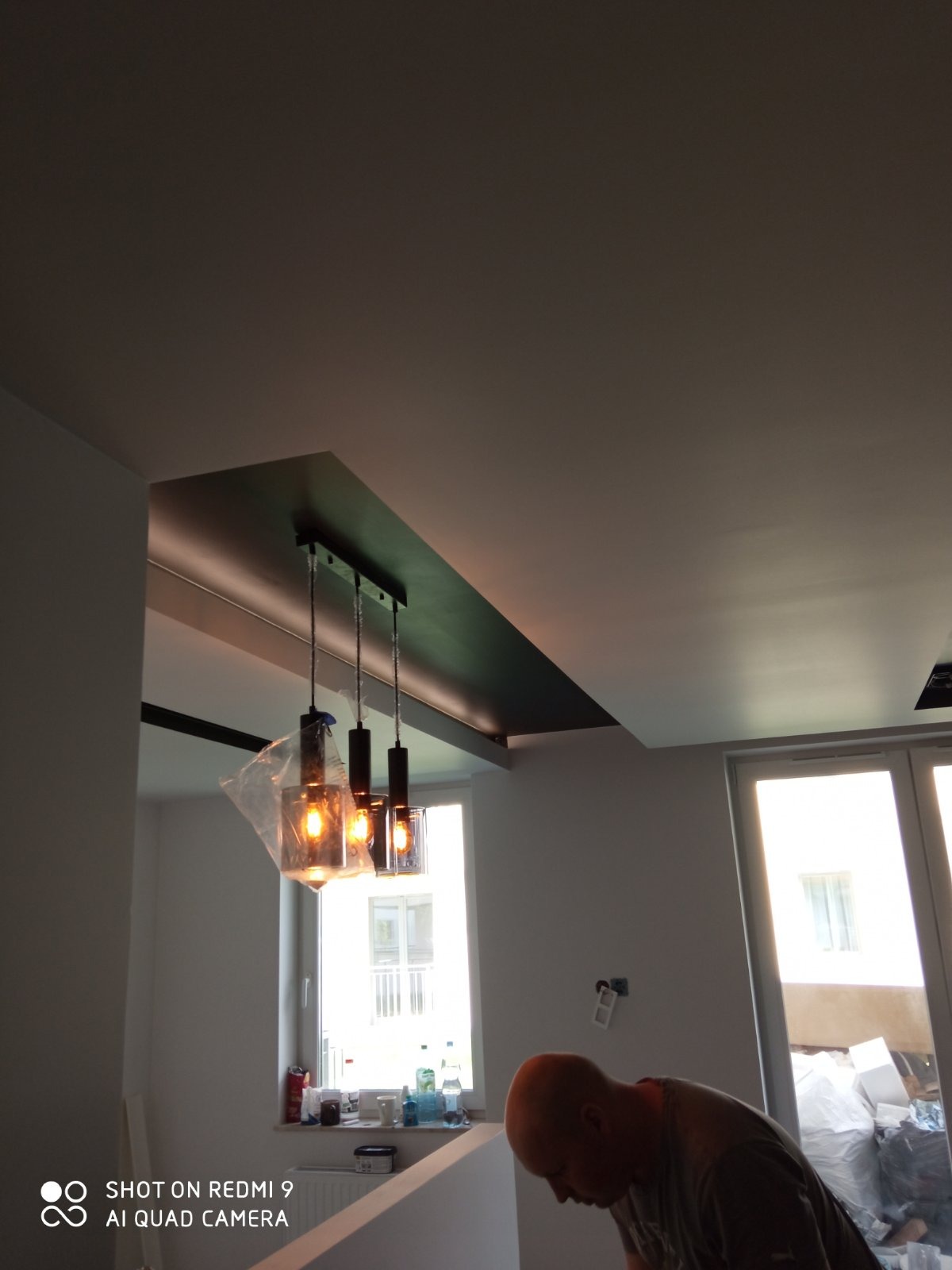 Montaż trzech wiszących lamp z żarówkami Edisona w nowym mieszkaniu, jedna z lamp jeszcze w folii ochronnej, widoczny fragment sufitu podwieszanego i okna.