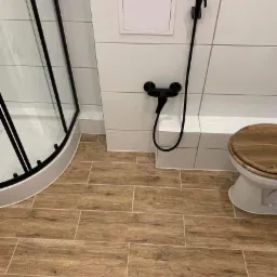 Nowoczesna łazienka z drewnopodobnymi płytkami podłogowymi, białymi płytkami ściennymi, czarną armaturą (bidetta i bateria) oraz częściowo widoczną kabiną prysznicową z czarnymi profilami i deską...