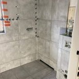 Marcin Rosłaniec - Wnętrze łazienki w trakcie układania szarych, wielkoformatowych płytek. Widoczne krzyżyki dystansowe i poziomica przy oknie. Częściowo ułożona podłoga, ściany w trakcie prac.