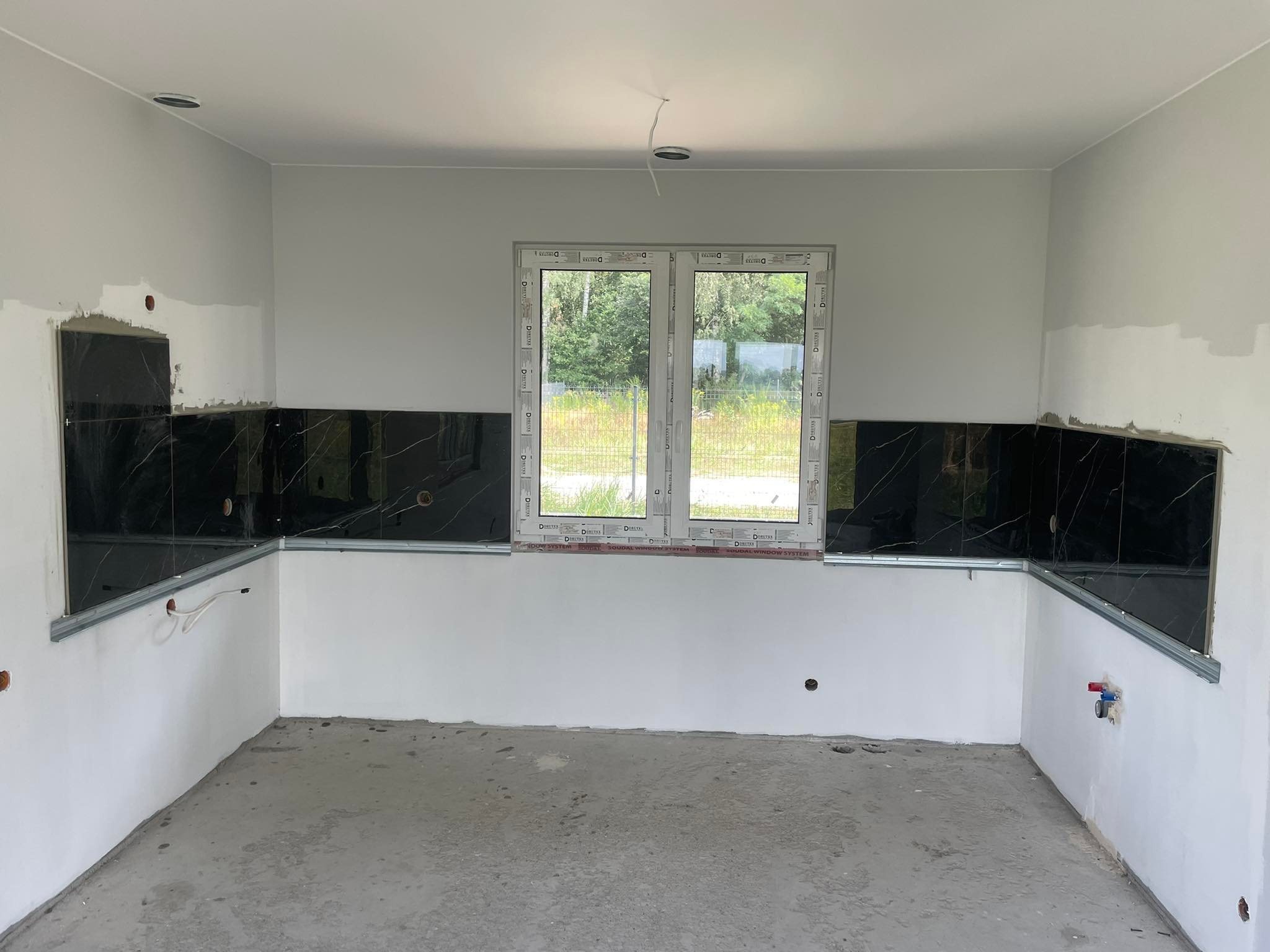 Wnętrze kuchni w trakcie układania czarnych płytek z marmurowym wzorem na ścianie, widoczne profile montażowe, surowy beton na podłodze i okno z widokiem na zieleń.