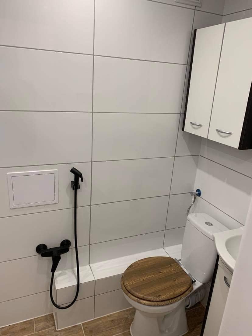 Nowoczesna łazienka z białymi, prostokątnymi płytkami, drewnianą deską sedesową, czarną baterią bidetową i białą szafką wiszącą z czarnymi bokami.