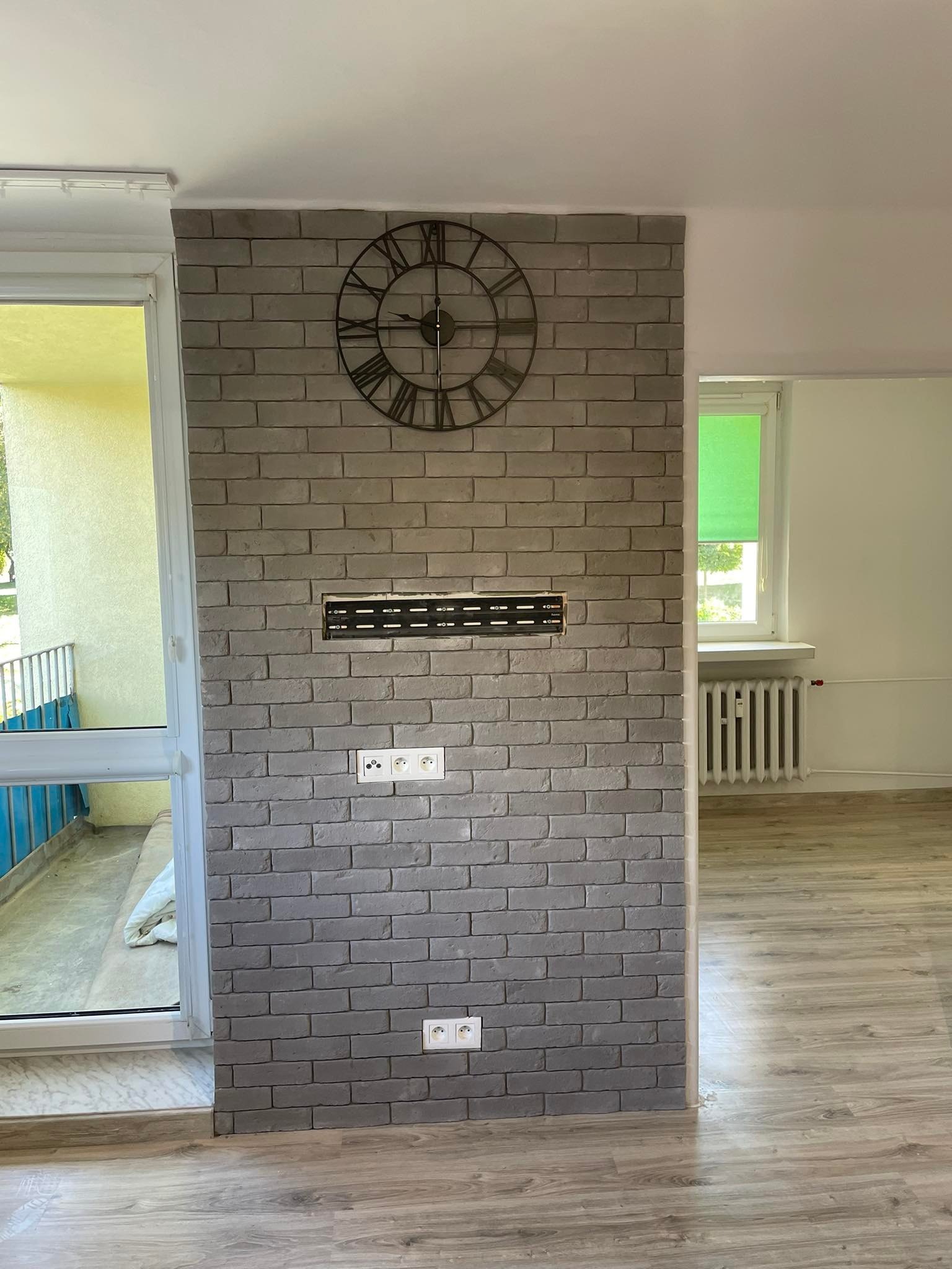 Ściana z szarej cegły dekoracyjnej z zamontowanym uchwytem do telewizora i zegarem w stylu vintage, widok na balkon i okno z zieloną roletą.