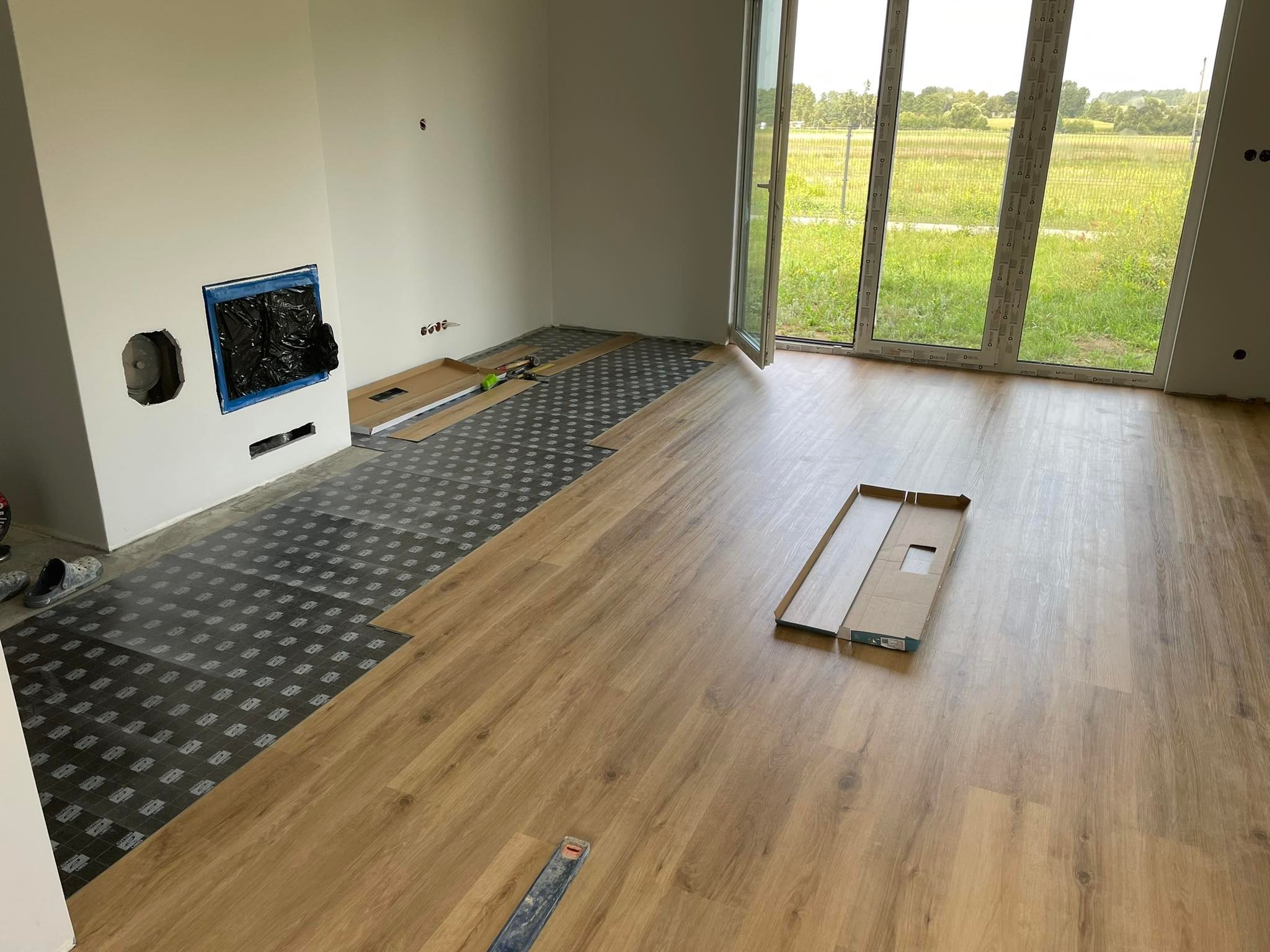 Układanie paneli podłogowych w jasnym pomieszczeniu z dużym oknem, widoczna podkładka izolacyjna i częściowo ułożone panele o wzorze drewna, zabezpieczony otwór kominkowy.