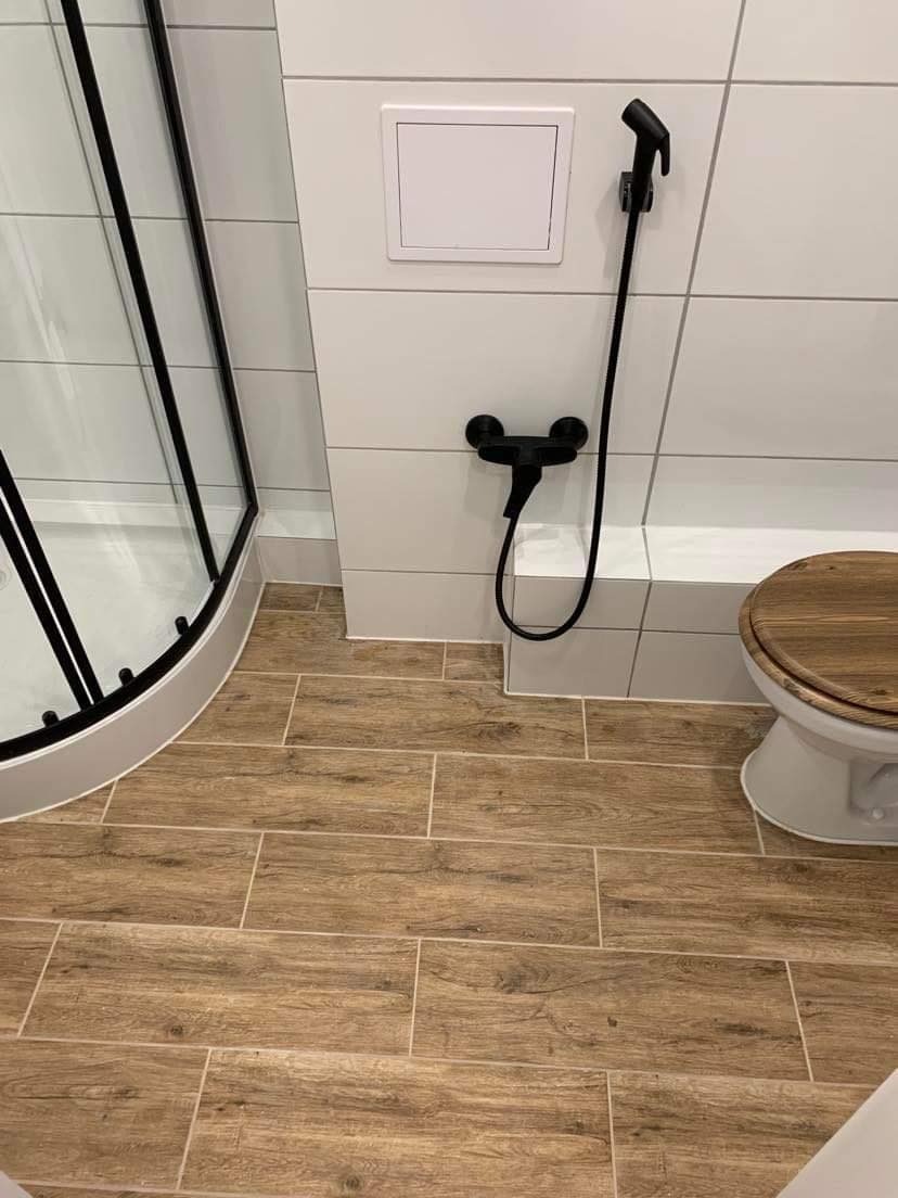 Nowoczesna łazienka z drewnopodobnymi płytkami podłogowymi, białymi płytkami ściennymi, czarną armaturą (bidetta i bateria) oraz częściowo widoczną kabiną prysznicową z czarnymi profilami i deską...