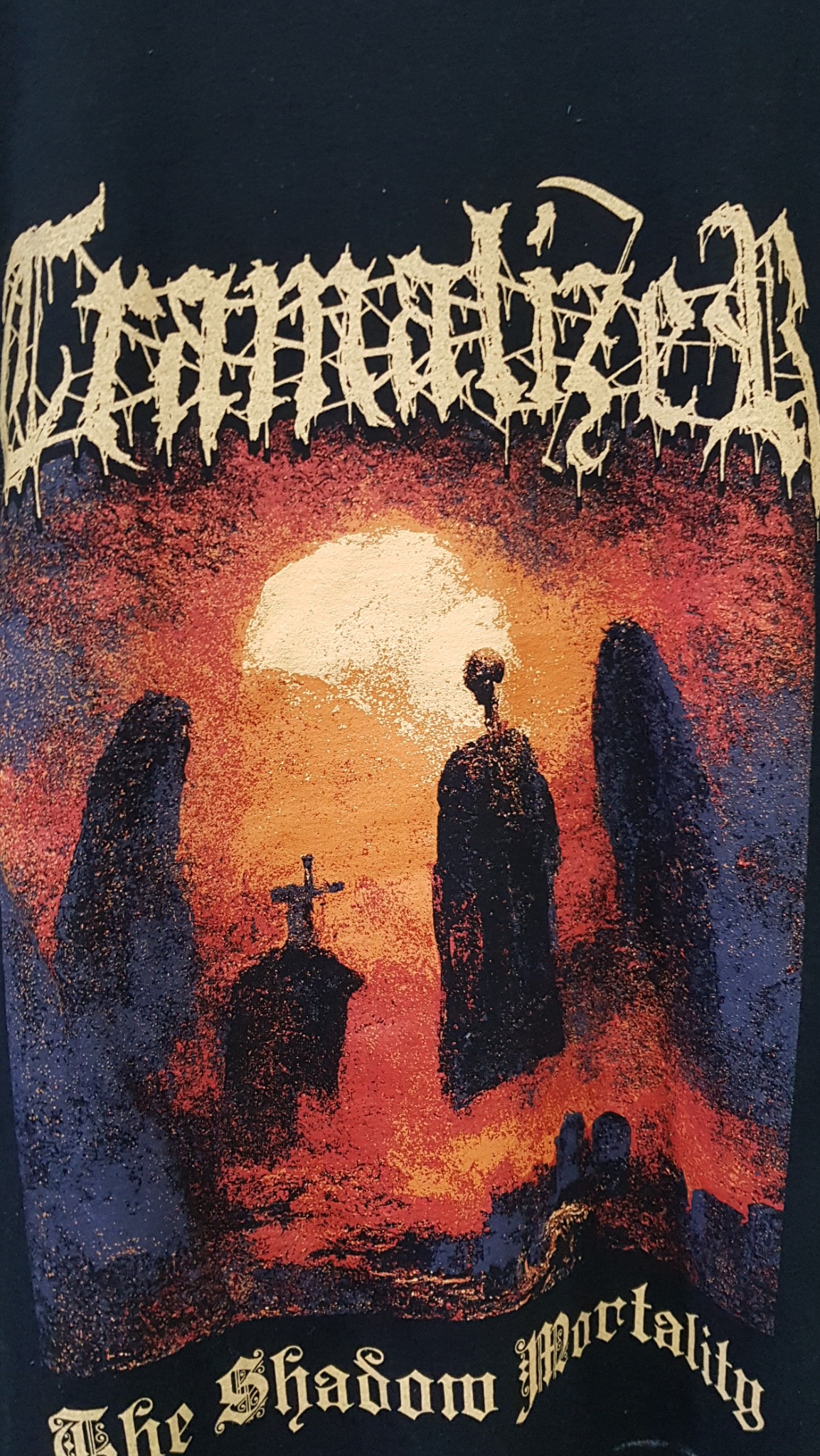 Czarna koszulka z nadrukiem w stylu death metal: cmentarz z nagrobkami, zachodzące słońce i tytuł albumu 'The Shadow Mortality'.