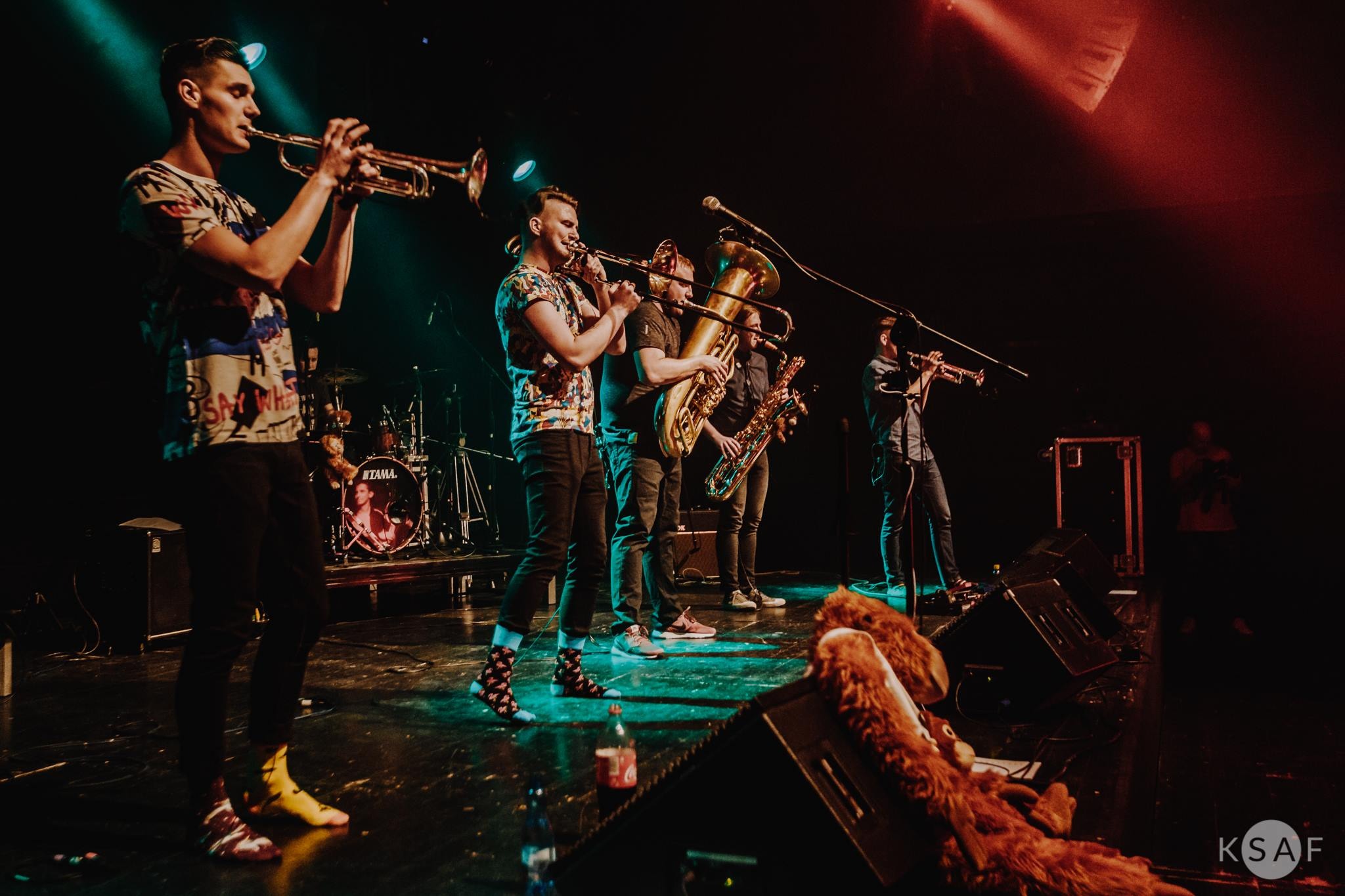 Zespół muzyczny grający na scenie: trąbka, puzon, tuba, saksofon, perkusja; wyróżniające się skarpetki w kropki i żółte buty trębacza z przodu.