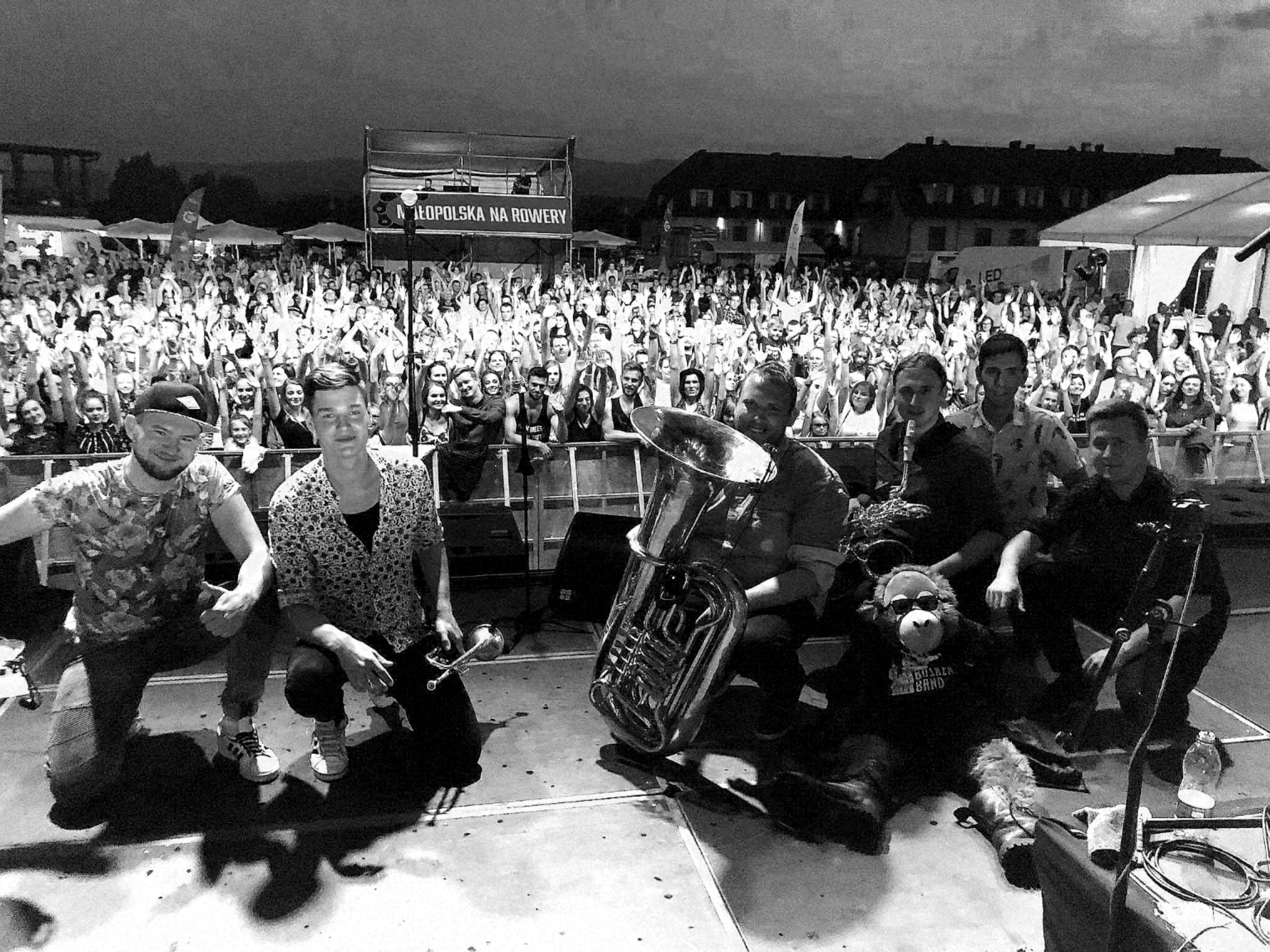 Czarno-białe zdjęcie zespołu muzycznego odpoczywającego na scenie po koncercie, w tle widoczny rozentuzjazmowany tłum fanów, na pierwszym planie tuba i pluszowy goryl w okularach.