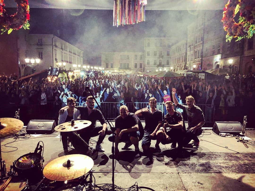Zespół muzyczny na scenie podczas koncertu plenerowego, w tle rozentuzjazmowany tłum publiczności i oświetlone budynki miejskie.