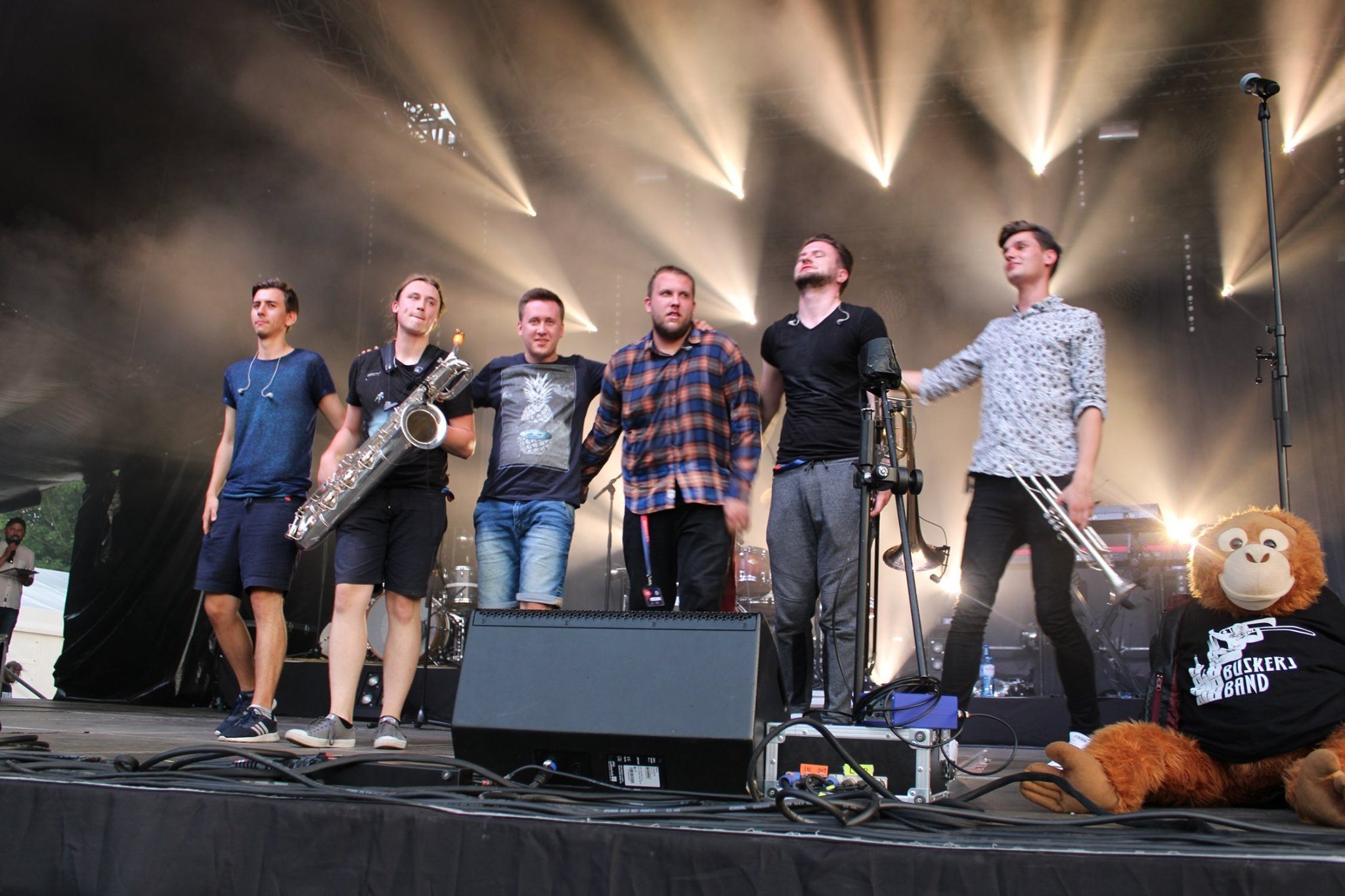 Zespół muzyczny na scenie podczas występu, w tle reflektory i maskotka małpy w koszulce z logo zespołu.