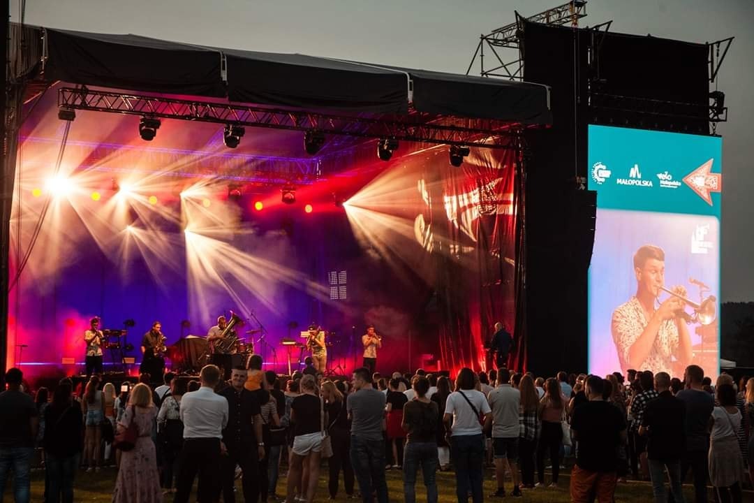 Scena plenerowa z zespołem muzycznym w trakcie występu, oświetlona reflektorami i z dużym ekranem LED po prawej stronie, widok z perspektywy publiczności.