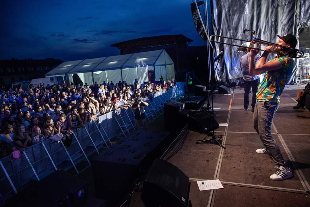Trombonista w koszuli w hawajskie wzory gra na scenie podczas koncertu plenerowego, widoczny tłum publiczności i sprzęt nagłośnieniowy.