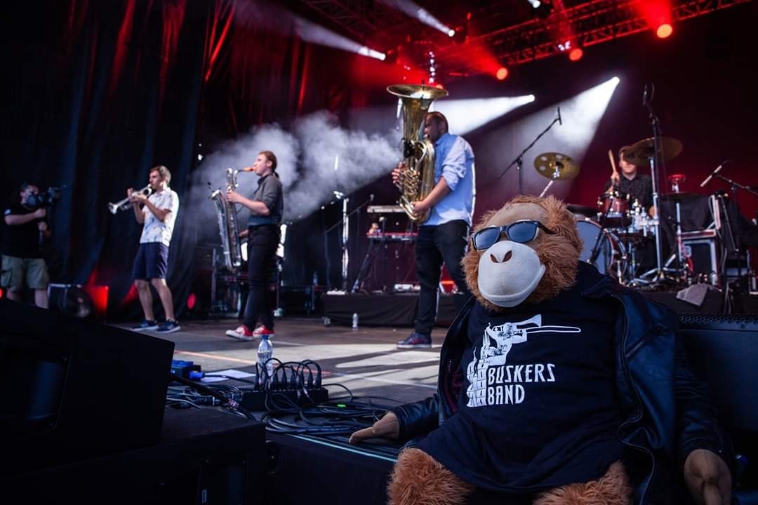 Pluszowy goryl w koszulce zespołu 'Busker Band' siedzi na skraju sceny podczas koncertu; w tle muzycy grający na trąbce, saksofonie, tubie i perkusji, oświetleni czerwonymi reflektorami.