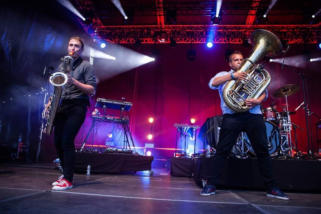 Zespół muzyczny na scenie: saksofon barytonowy i tuba na tle czerwonej kurtyny i świateł scenicznych.