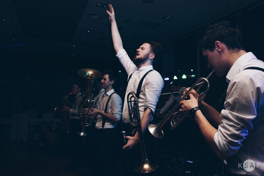 Zespół muzyczny w białych koszulach i szelkach grający na instrumentach dętych podczas występu na scenie, wokalista z podniesioną ręką.