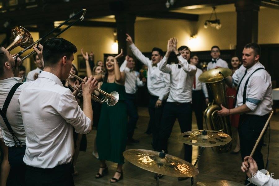 Zespół muzyczny grający na żywo podczas imprezy, widoczni trębacze, perkusja i tuba na tle tańczących gości.