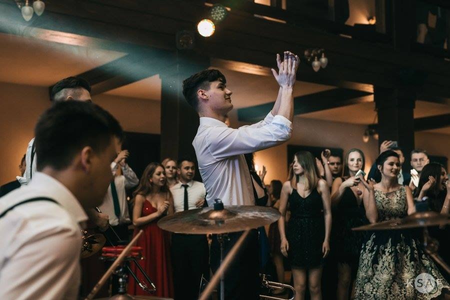 Zespół muzyczny na scenie podczas występu dla publiczności, widoczne instrumenty perkusyjne i oświetlenie sceniczne.