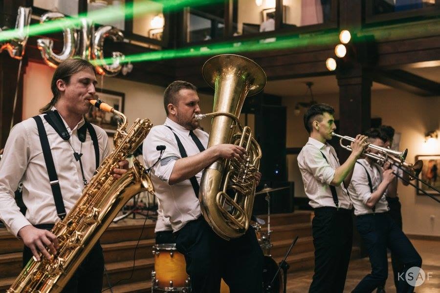 Zespół muzyczny w białych koszulach i czarnych spodniach z szelkami grający na saksofonie, tubie i trąbkach podczas występu na scenie z dekoracjami balonowymi.