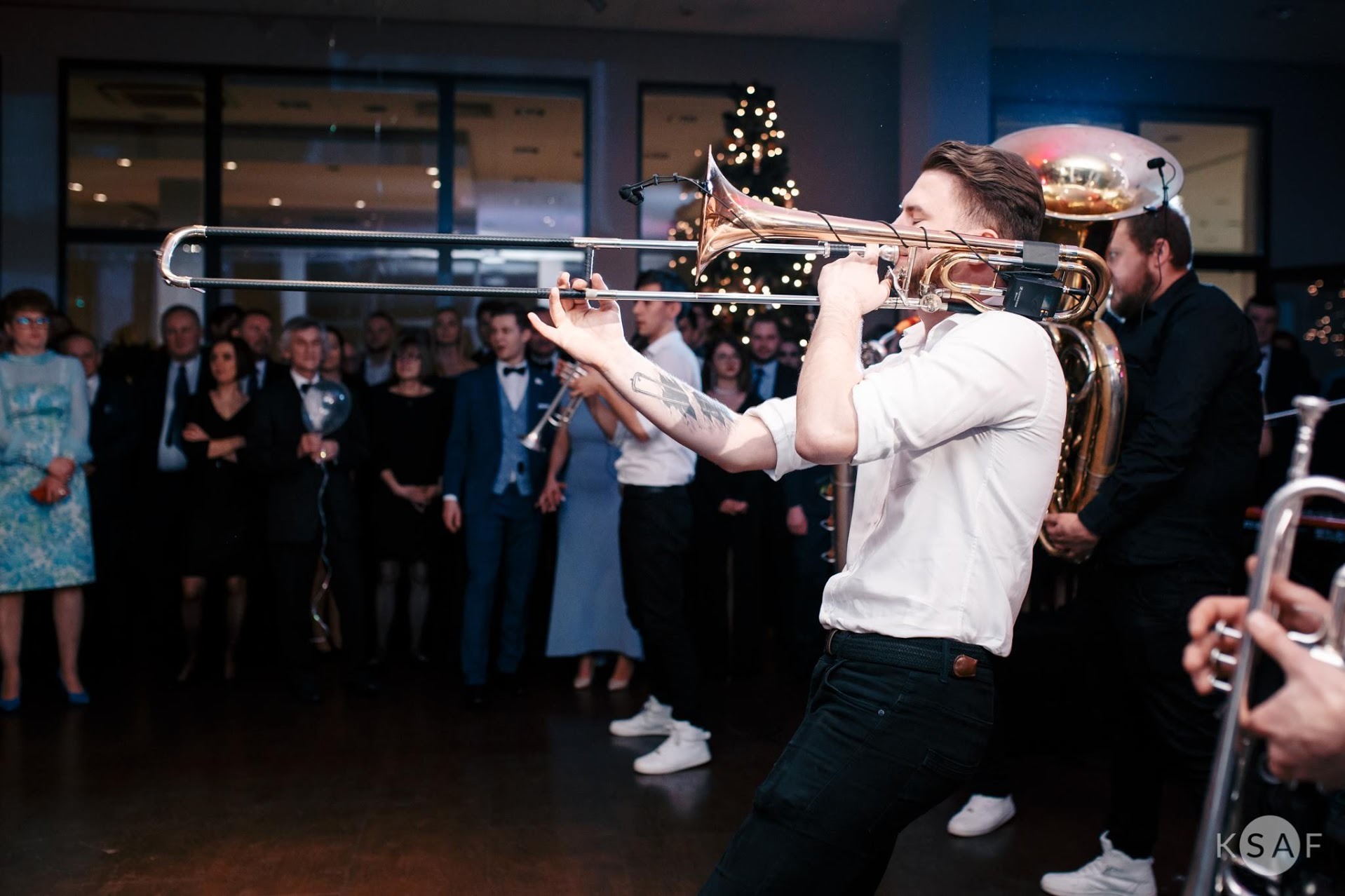 Muzyk grający na puzonie na tle publiczności podczas wydarzenia, widoczna tuba i choinka w tle.