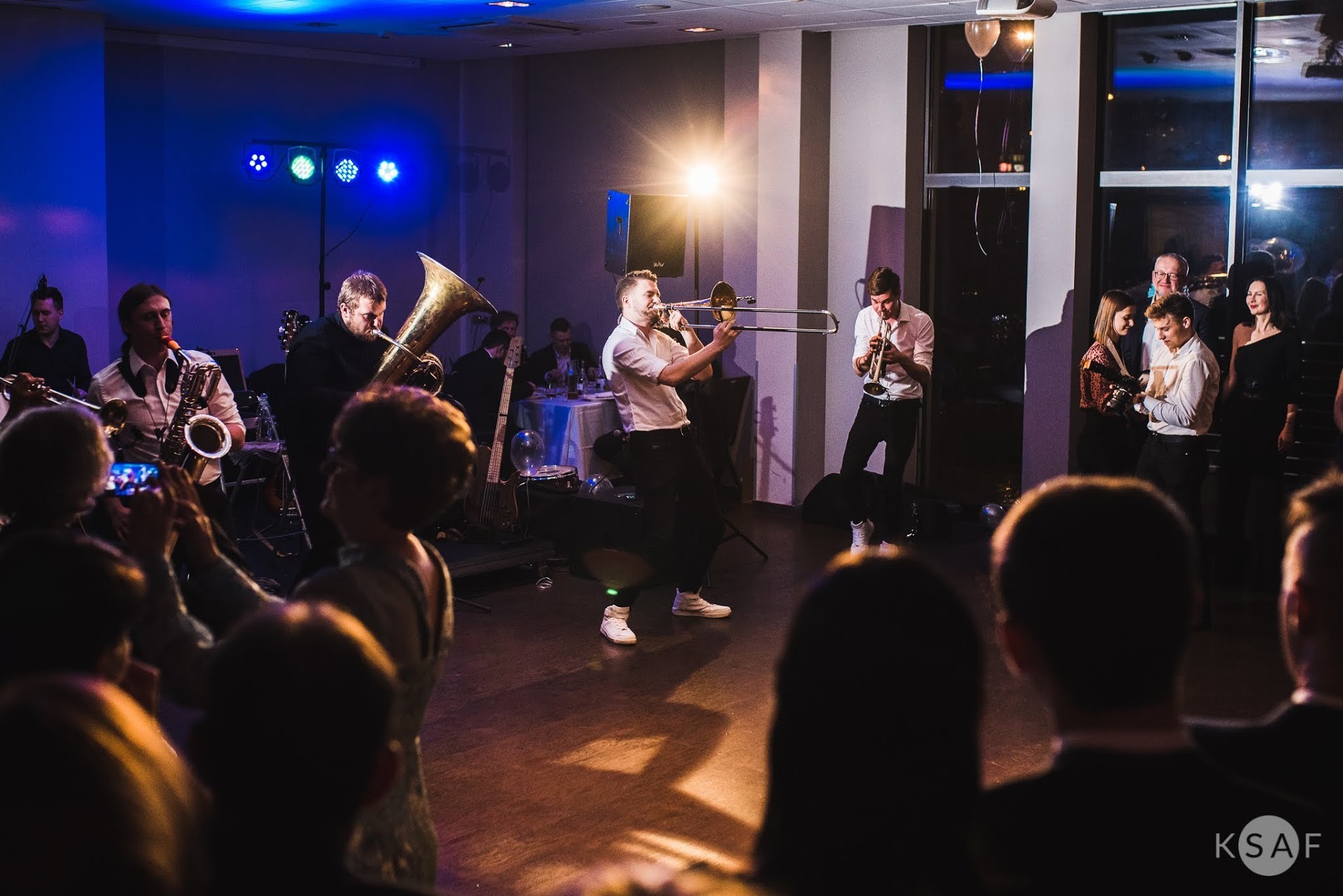 Zespół muzyczny gra na instrumentach dętych podczas imprezy, oświetlenie sceniczne w kolorze niebieskim i białym, widownia obserwuje występ.