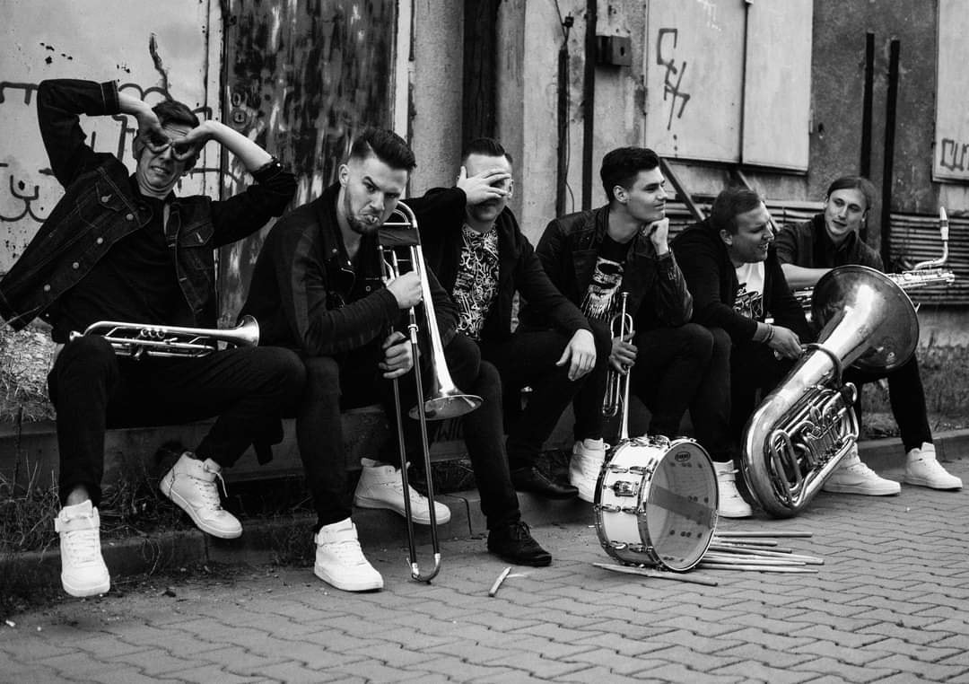 Czarno-białe zdjęcie zespołu muzycznego siedzącego na chodniku z instrumentami: trąbki, puzon, tuba i bęben. Jeden z muzyków robi gest dłońmi nad głową, imitując rogi.