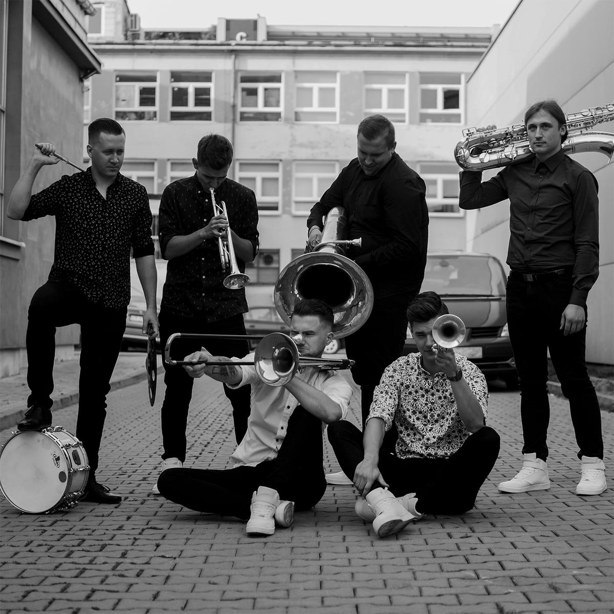 Czarno-białe zdjęcie zespołu muzycznego w nieformalnym ustawieniu: trębacz, puzonista siedzący na ziemi, saksofonista, perkusista z pałeczką opartą o bęben i tubista, na tle miejskiej uliczki...